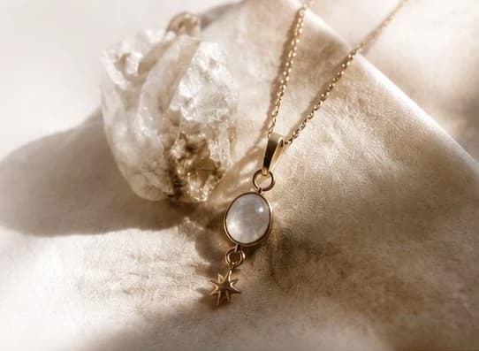 A moonstone pendant and a small gold star charm on a fine gold chain, beside a raw quartz crystal on cream linen